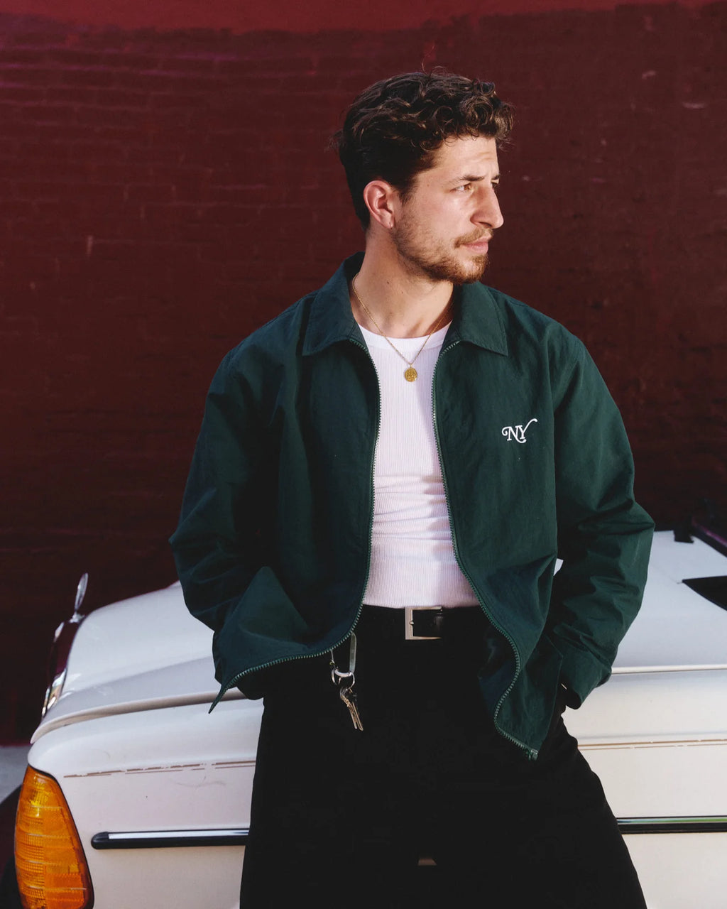 A man wearing a dark green bomber jacket over a white t-shirt and black pants, standing next to a vintage white car against a dark red brick wall.