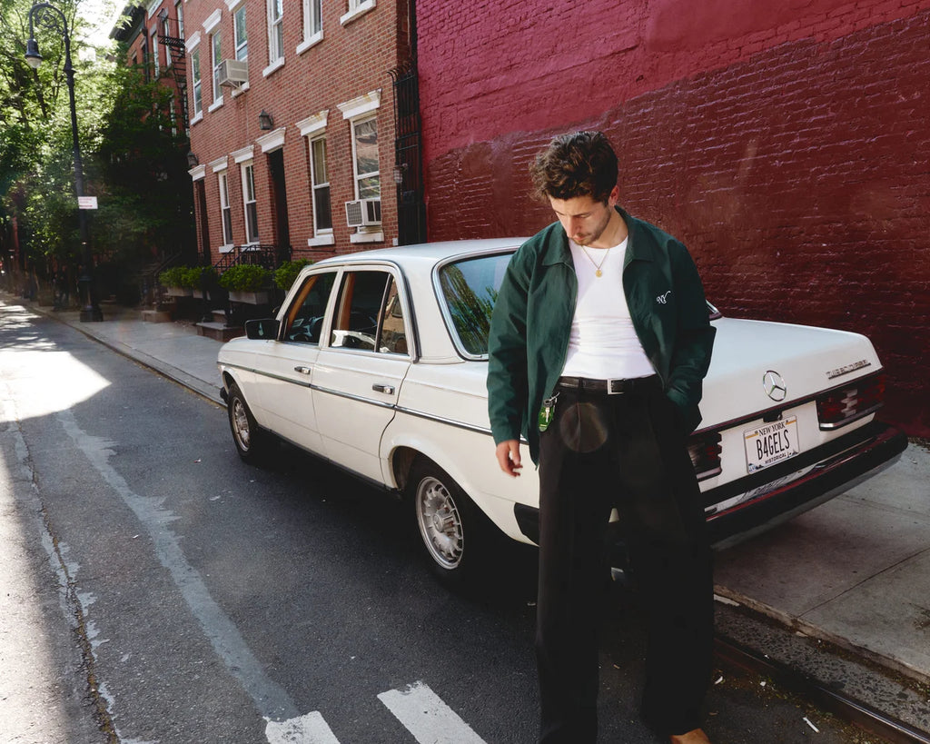 A man wearing a dark green bomber jacket, white t-shirt, and black wide-leg trousers stands next to a white vintage Mercedes-Benz sedan parked on a city street with a red brick wall in the background.
