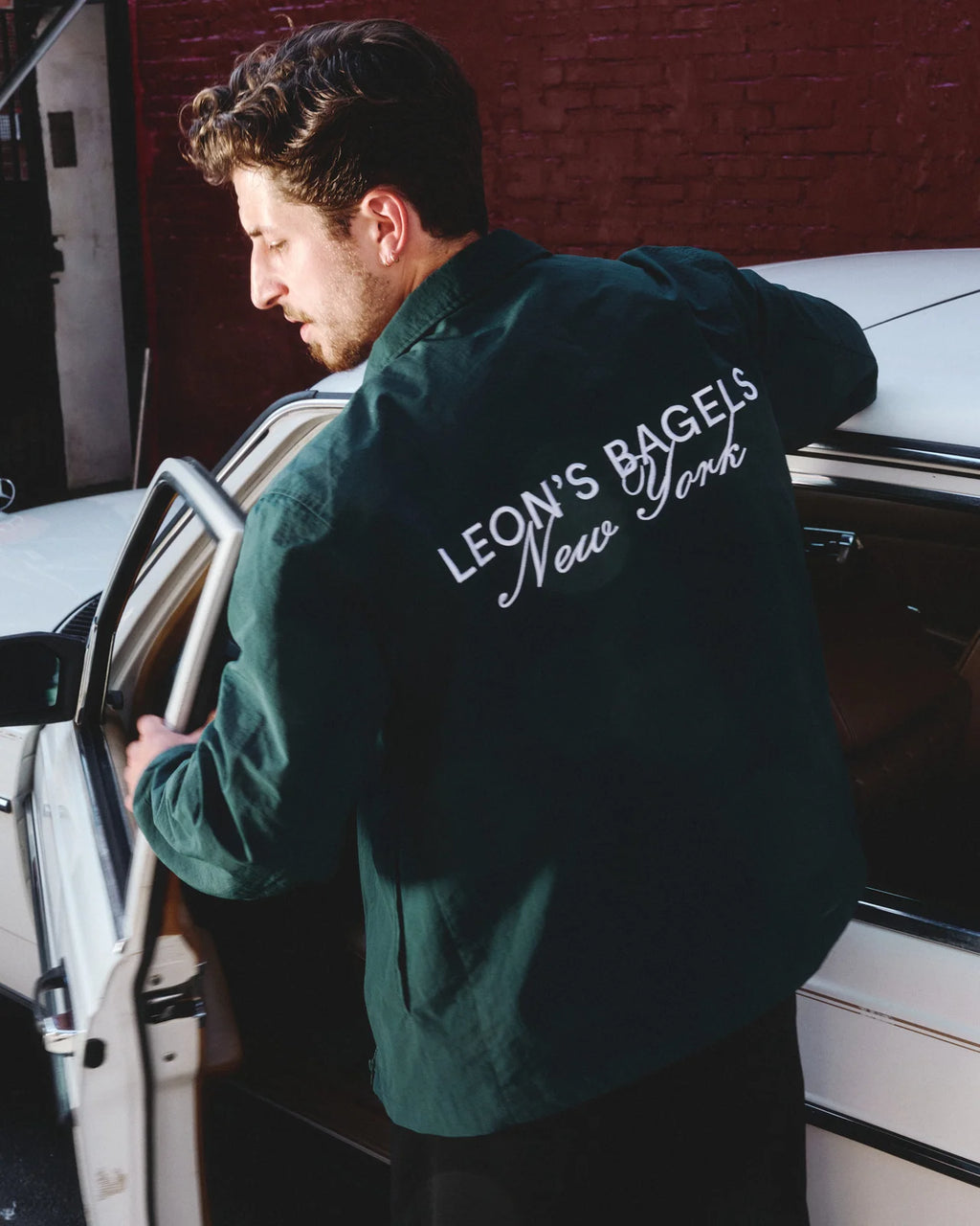 A man wearing a dark green jacket with 'LEON'S BAGELS New York' printed in white on the back, standing next to a white car in front of a red brick wall.