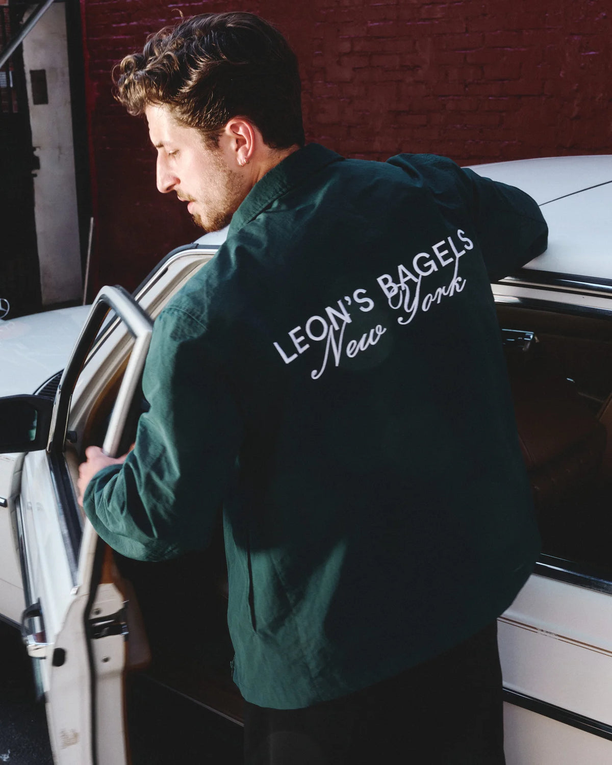 A man wearing a dark green jacket with 'LEON'S BAGELS New York' printed in white on the back, standing next to a white car in front of a red brick wall.