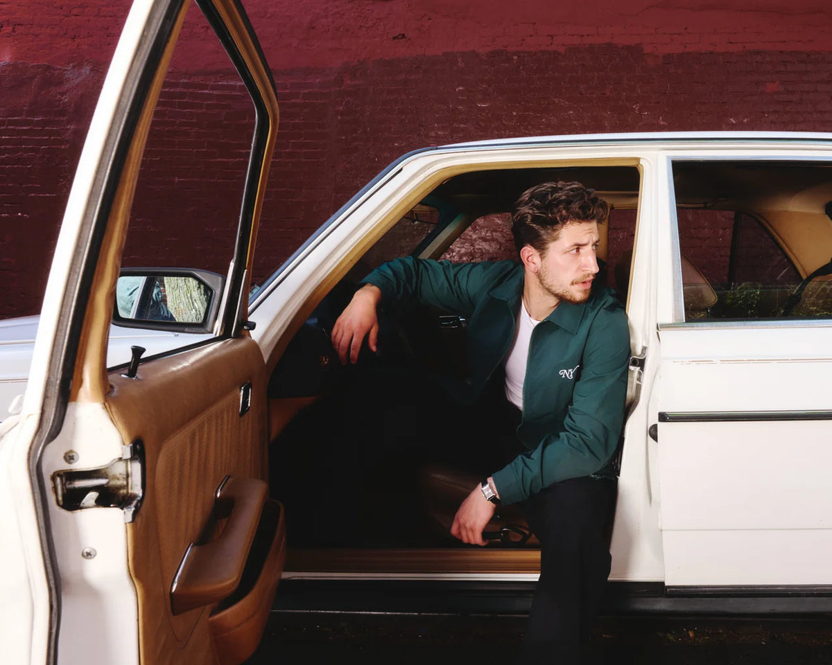 A man wearing a dark green bomber jacket and black pants sits in the open door of a vintage white car with a brown leather interior, set against a red brick wall.