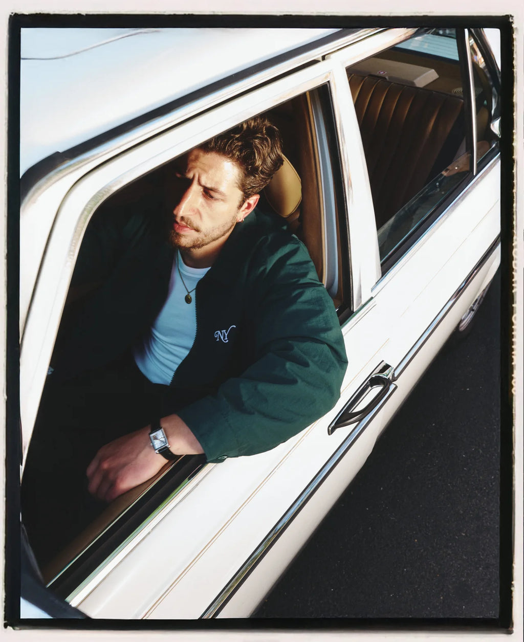 A man leans out of a white car window wearing a dark green bomber jacket over a light blue t-shirt. The photo has a vintage, slightly tilted perspective with a black border.