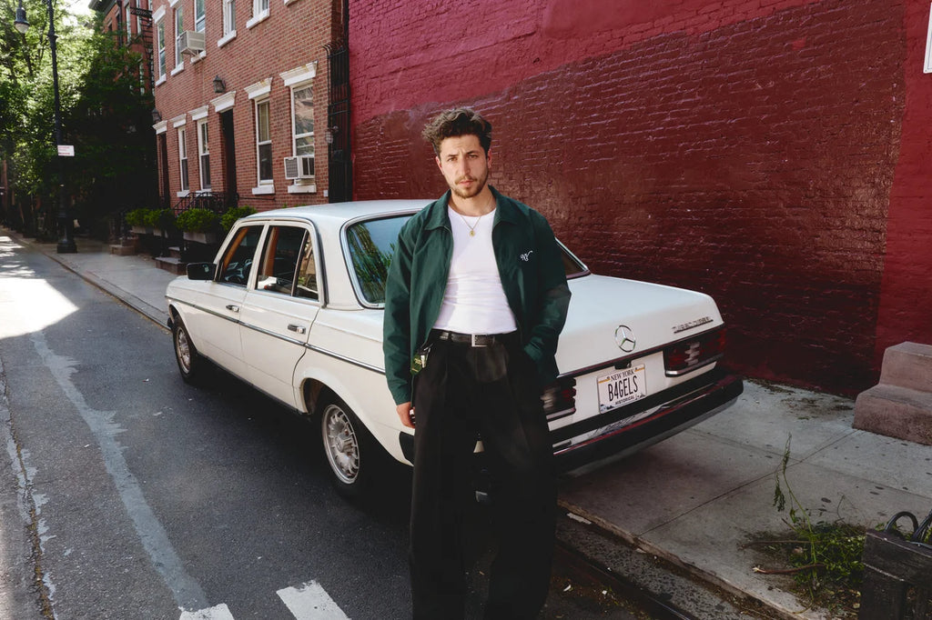 A man stands on a city sidewalk next to a white vintage car, wearing a dark green bomber jacket over a white t-shirt and black pants, with a red brick wall in the background.