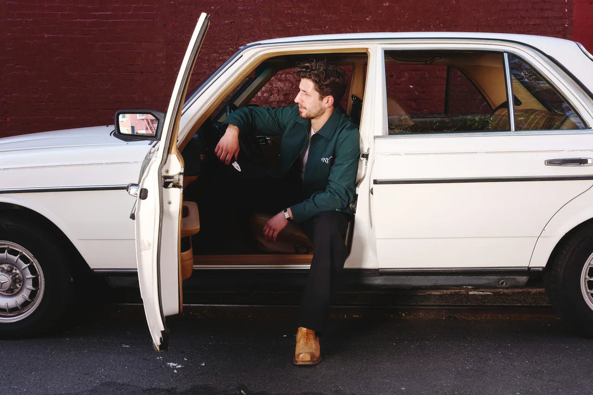 A man in a dark green jacket and black trousers sits in the open driver's side door of a white vintage car, with a dark red brick wall in the background.