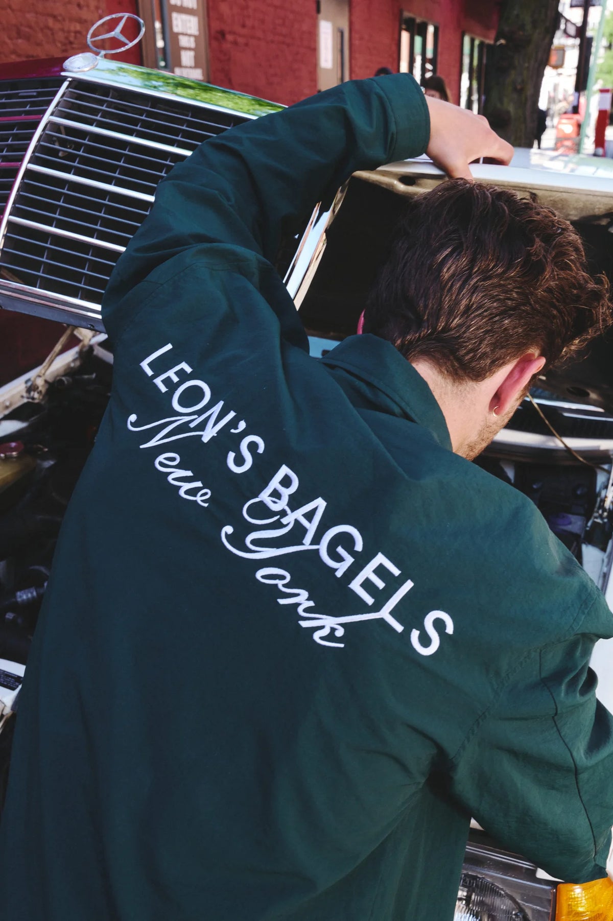 A man seen from behind wearing a dark green jacket with 'LEON'S BAGELS New York' printed in white script across the back, leaning over the front of a vintage car in an outdoor street setting.