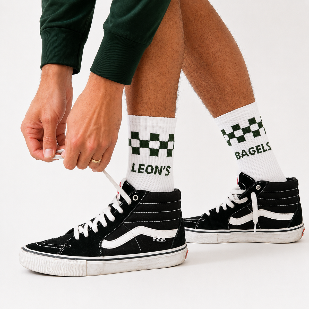 A person wearing black high-top sneakers with white socks featuring a green and white checkerboard pattern and the names 'LEON'S' and 'BAGELS' printed on them. The person is tying the laces of the left shoe against a clean white background.