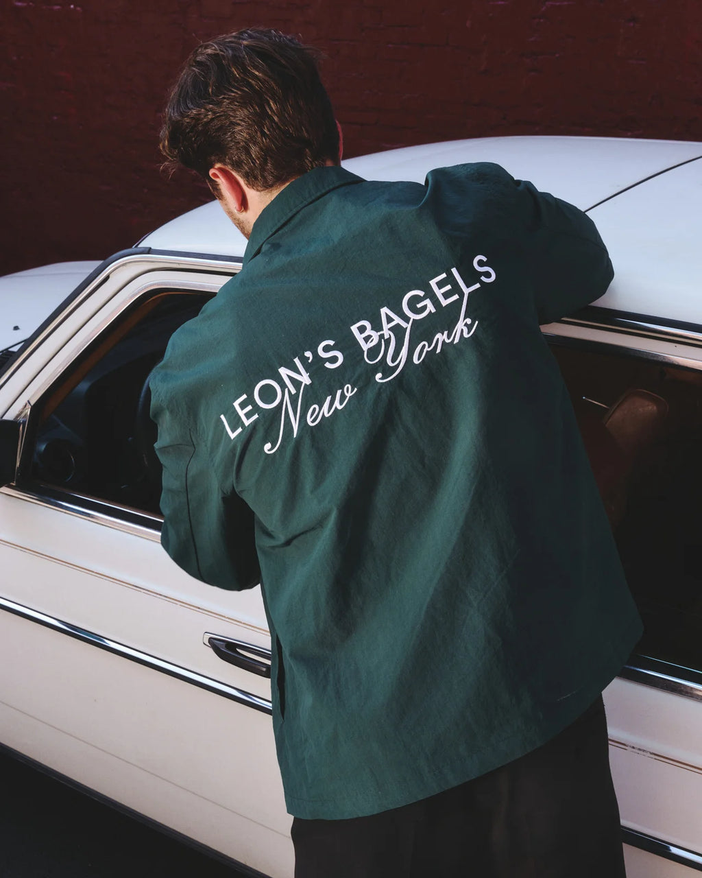 A man wearing a dark green overshirt with 'LEON'S BAGELS New York' printed in white script across the back, leaning against a white vintage car in front of a red brick wall.