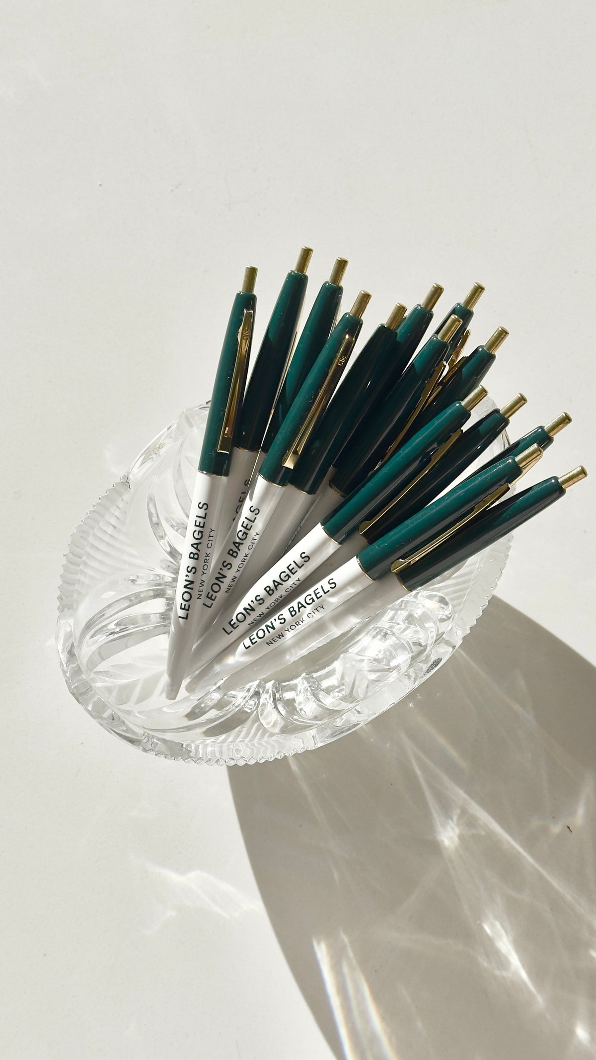 A set of Leon & Bagels 45mm ballpoint pens with dark teal barrels, silver metal accents, and gold tips, arranged in a clear glass dish on a white surface.