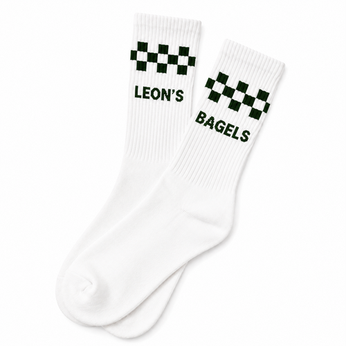 A pair of white crew socks with a green checkered stripe and the words 'LEON'S BAGELS' printed in green near the cuff, shown on a plain white background.